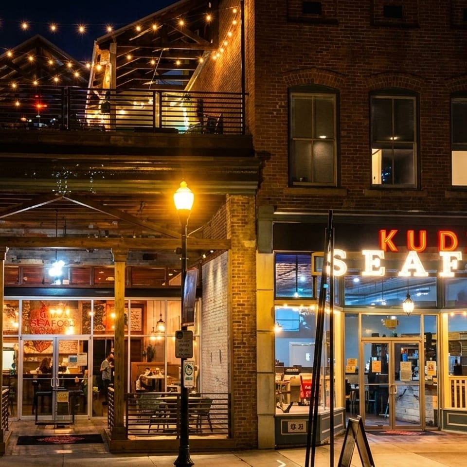 Exterior night view of Kudzu Seafood restaurant with illuminated signs, outdoor seating, and string lights.