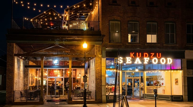 Exterior night view of Kudzu Seafood restaurant with illuminated signs, outdoor seating, and string lights.