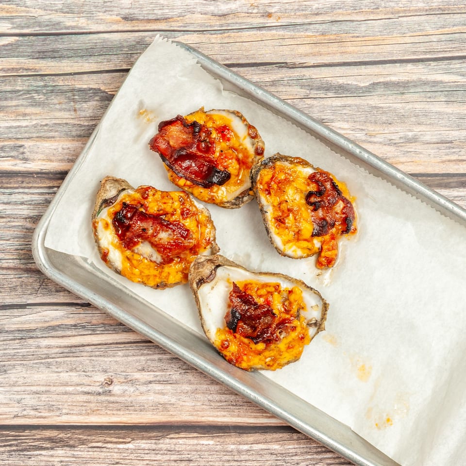 Four baked oysters topped with melted cheese and crispy bacon on a metal tray with parchment paper.