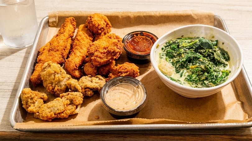 A tray of fried chicken, fried oysters, two dipping sauces, and a bowl of creamed spinach.