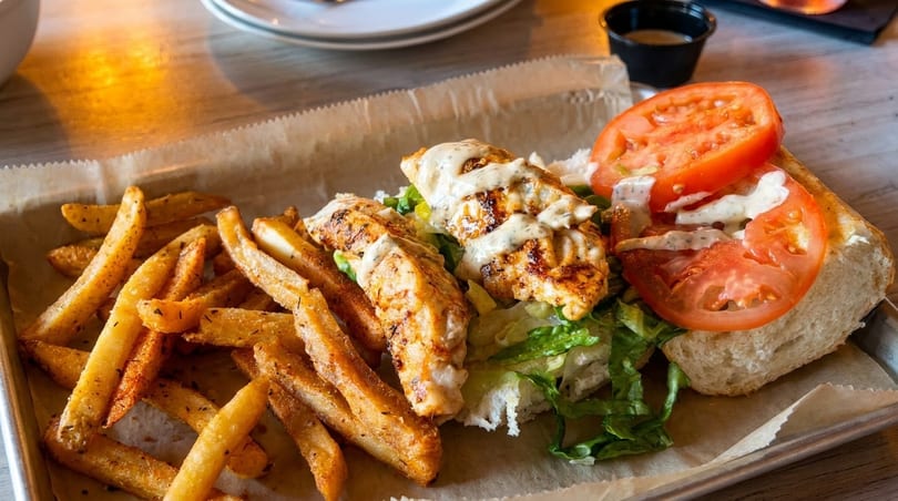 Grilled chicken po'boy with lettuce, tomato, and sauce, served with seasoned fries on a metal tray.