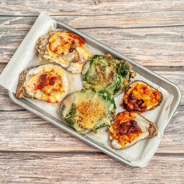 Six baked oysters with various toppings, including cheese, spinach, and red peppers, on a metal tray.