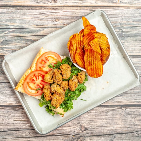 Fried oyster po'boy sandwich with lettuce, tomato, and a side of waffle-cut chips on a tray.