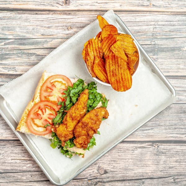 Fried chicken sandwich with lettuce, tomato, and a side of waffle chips on a metal tray.