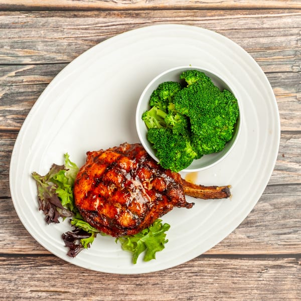 Glazed pork chop with a side of steamed broccoli and mixed greens on a white plate.