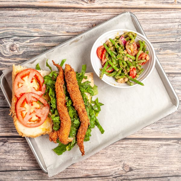 Fried chicken sandwich with lettuce and tomato, served with a side of green bean salad.