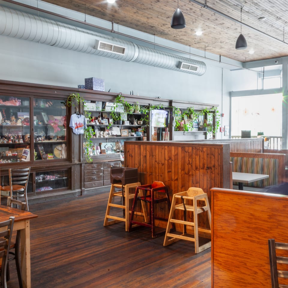 Restaurant interior with dark wood floors, display cases, high chairs, and booth seating near bright windows.
