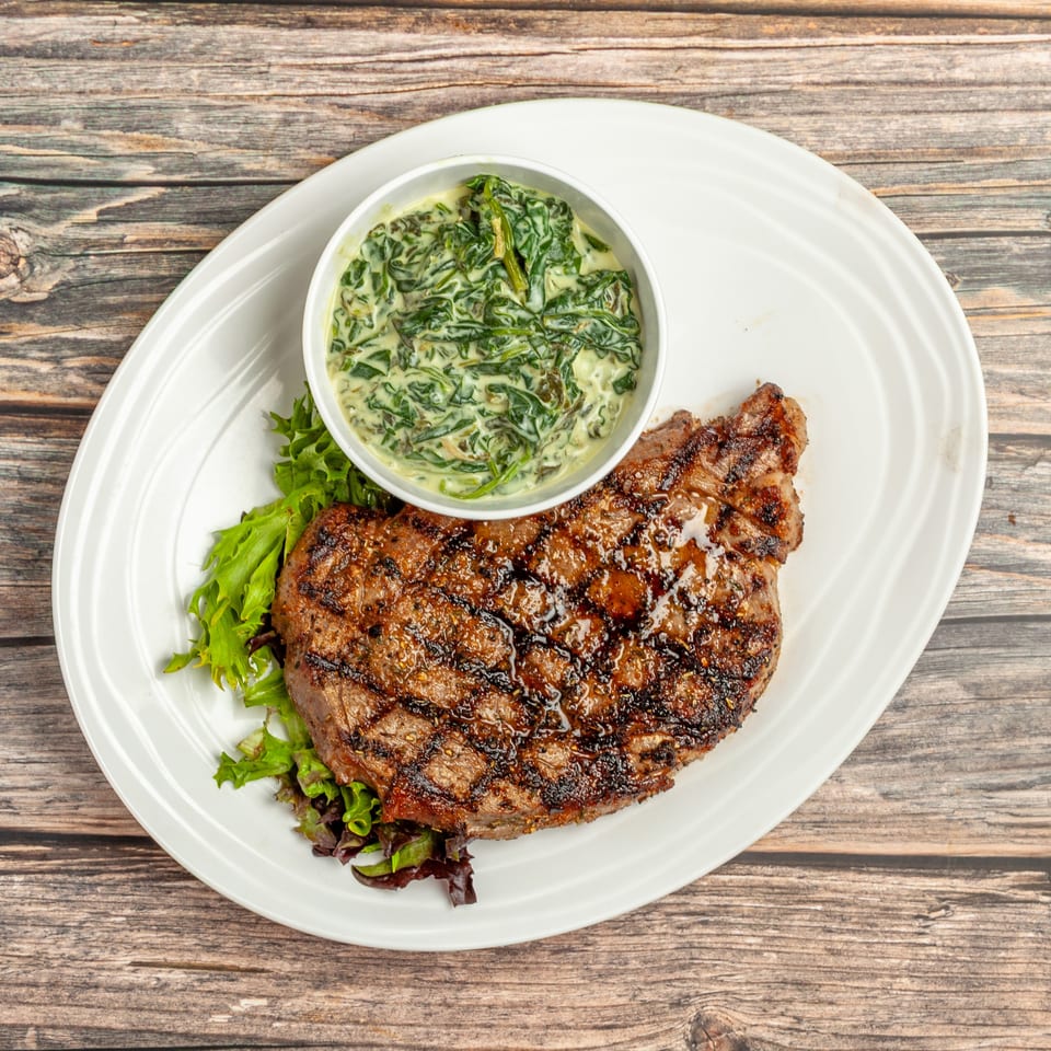 Grilled steak with grill marks, served with creamed spinach and a side salad on a white plate.